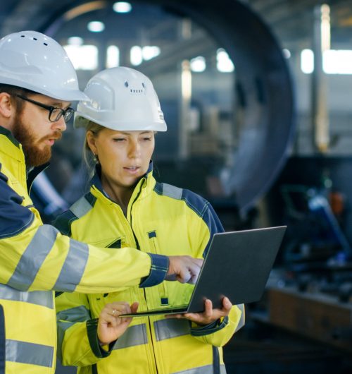 Male and Female Industrial Engineers in Hard Hats Discuss New Pr Male and Female Industrial Engineers in Hard Hats Discuss New Project while Using Laptop. They Make Showing Gestures.They Work in a Heavy Industry Manufacturing Factory.